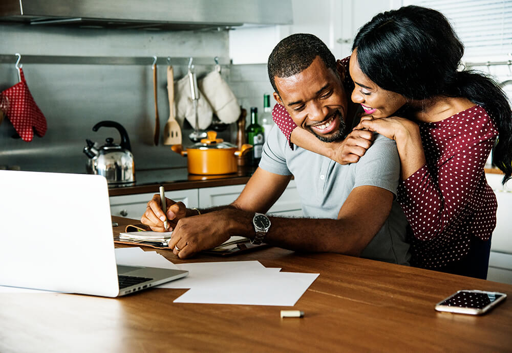 A couple at a kitchen table with a laptop, discussing and collaborating on their tasks