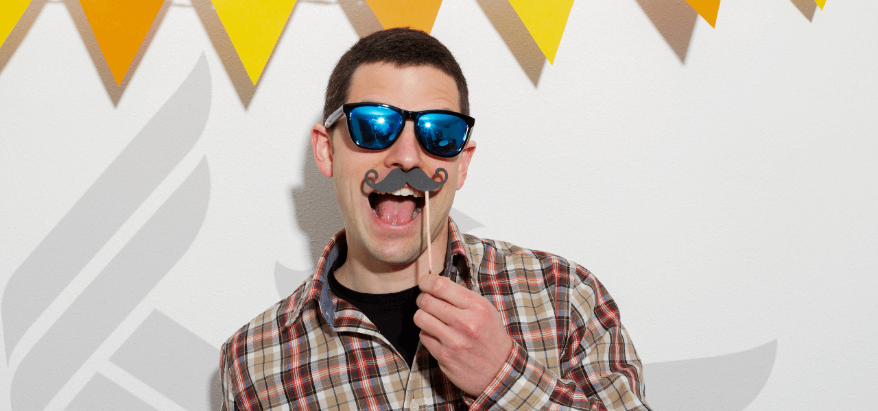 A University of Phoenix Alumnus wearing sunglasses and a fake moustache, posing for a picture