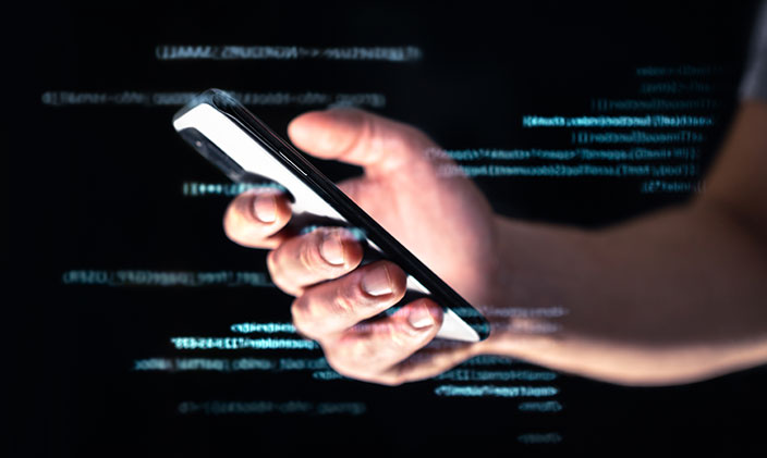 Person's arm and hand holding a smart phone with code in the background to represent spyware