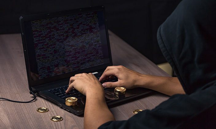 A person sitting in a darkened room uses a laptop with gold coins scattered nearby to signify cryptojacking