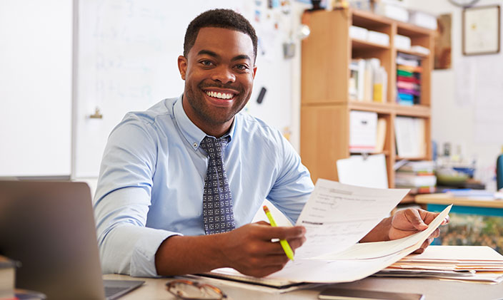 A smiling teacher reviews class curriculum documents at his desk