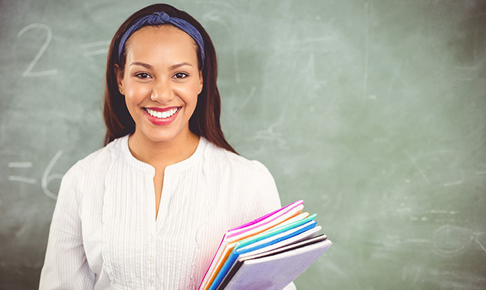 A photo of a smiling african-american female educator holding folders in front of a blackboard to signify the pursuit of education fellowships.