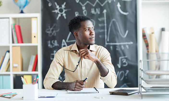 Male secondary education teacher sitting at his desk in a classroom in front of a chalkboard