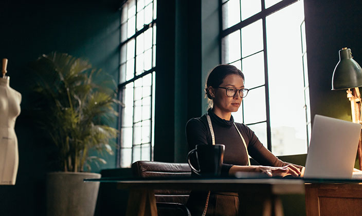 Female fashion entrepreneur types on a laptop while wearing measuring tape around her neck, searching online for degrees for entrepreneurs
