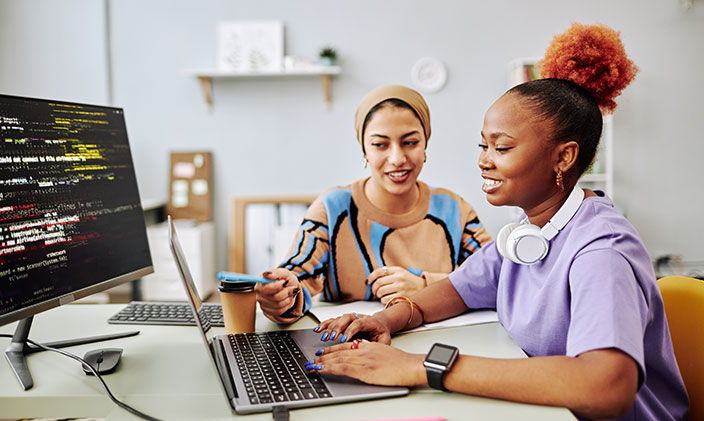 Two female interns reviewing computer code on a laptop and monitor screen as part of their tech internship.