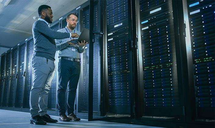 Two technology specialists overlooking work on laptops, standing in front of a massive computer structure behind glass walls
