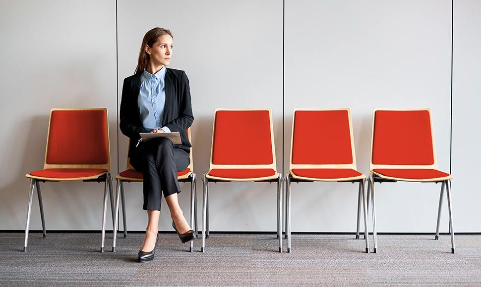 A job seeker sits alone, cross-legged in a row of five chairs, anxiously awaiting an interview.