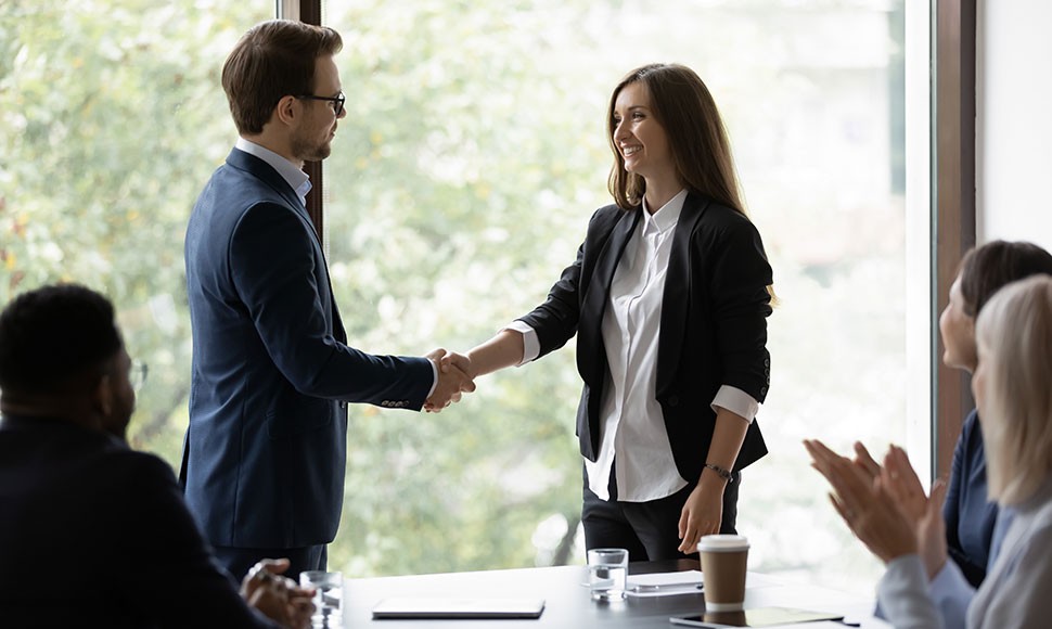 Man and woman shake hands in front of a conference table of clapping people