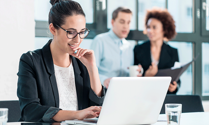 Introverted professional smiles faintly at her laptop while gossipy coworkers chat in the background