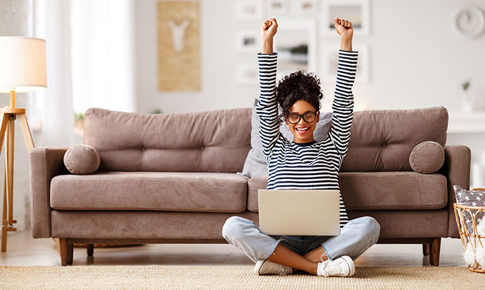 Professional woman gleefully raising both hands while sitting in front of her couch with a laptop in her lap