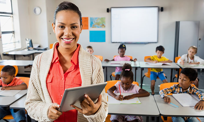 Elementary teacher standing in front of classroom holding iPad and smiling with students hard at work in the background