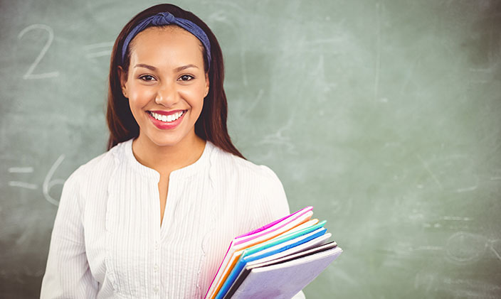 smiling-female-teacher-holding-colorful-folders-in-front-of-chalkboard