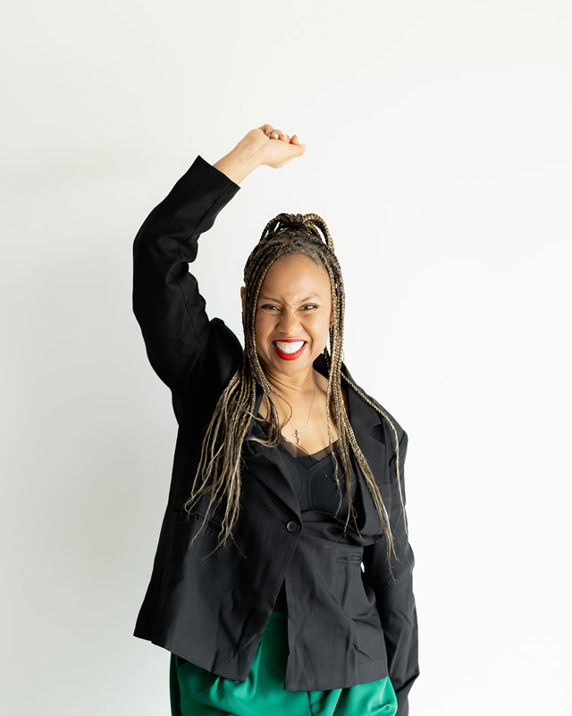 Freda Richards raising a fist and smiling in a confident pose, wearing a black blazer and green pants.