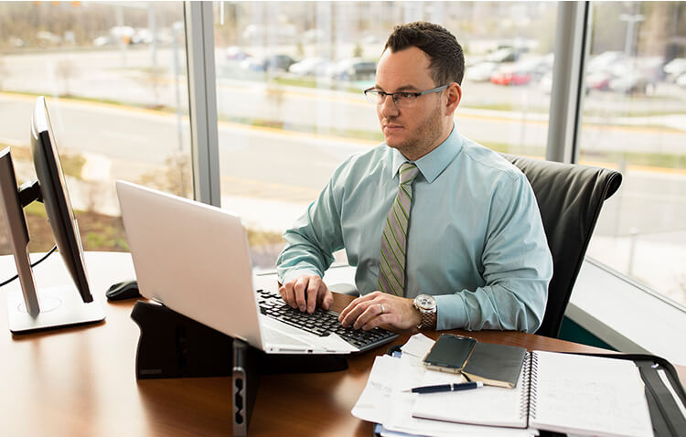 An financial analyst sits at a desk, using a laptop, in front of a bank of windows