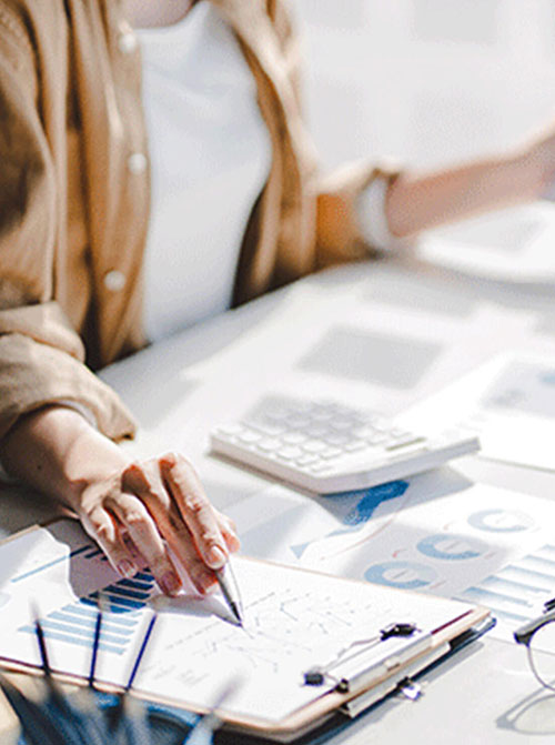 A financial analyst, seated at a desk covered with papers showing charts, points to something on a clipboard