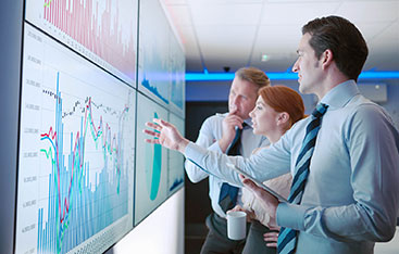 Three professionally dressed colleagues look at a wall of charts and graphs together