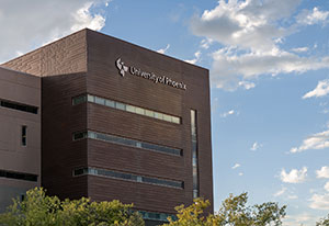 Link to an image of the University of Phoenix building in front of a blue sky with light clouds, with the building in the lower left portion of the frame