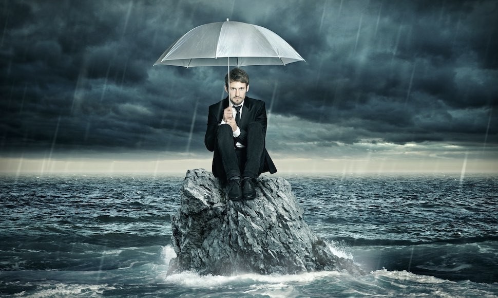Man wearing suit and tie, holding umbrella and sitting on a rock in a stormy sea.
