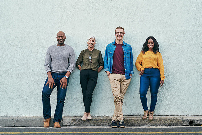 Four people of different ages, genders, and racial backgrounds leaning against a wall together