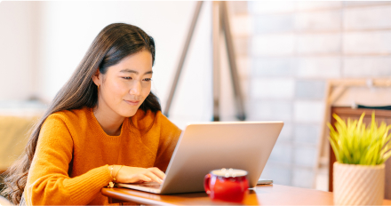 A woman in an orange sweater sitting at a table, focused on using a laptop computer. 