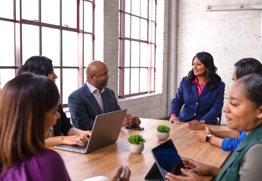 Several people sit around a conference table with laptops and tablets, talking in a bright room with large windows.