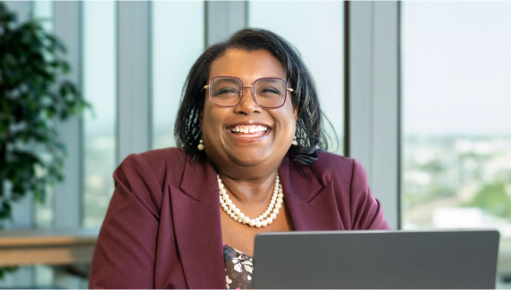 A confident, smiling alumna sits at a desk in a modern office, symbolizing University of Phoenix MBA education is relevant and practical.