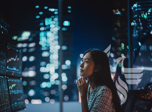 Woman looking thoughtfully at computer monitors backlit by a brightly lit city skyline