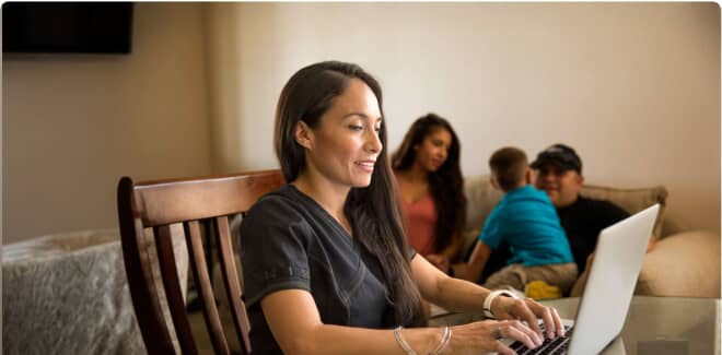A student in a black shirt sitting at a desk and smiling while working on their laptop with their family in the background
