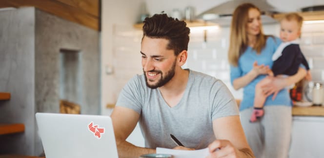 A University of Phoenix student works on his laptop at the kitchen table with his wife and baby nearby