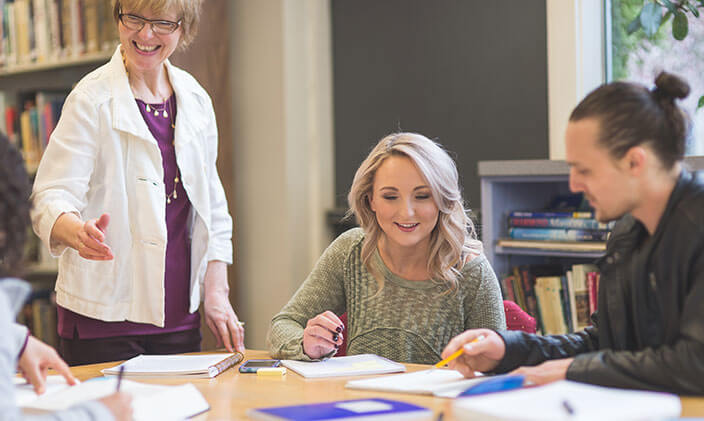 An instructor and two adult students gather around a table in a library to discuss a project