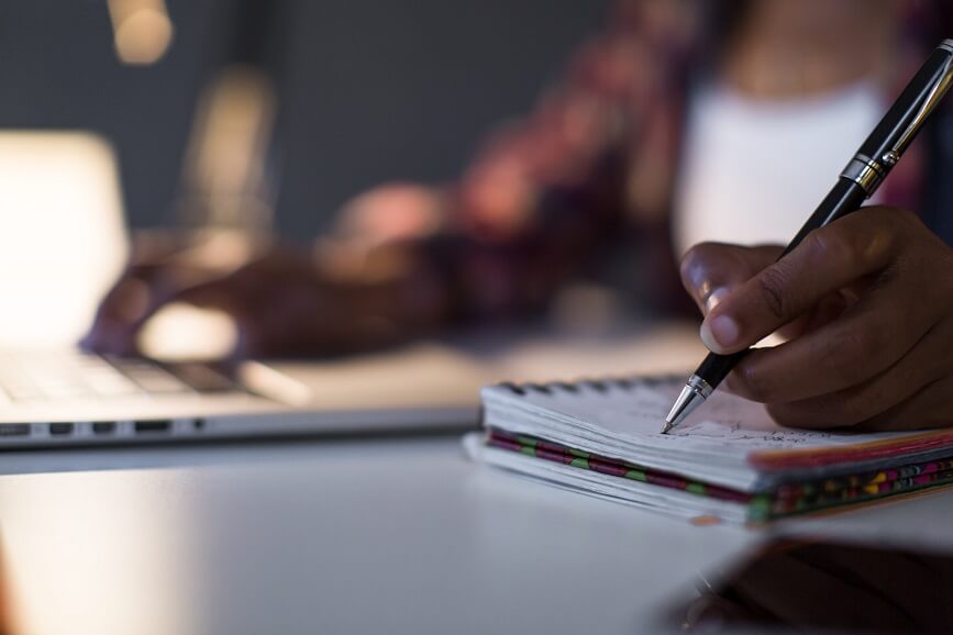 A person writing in a notebook while referencing information on a laptop