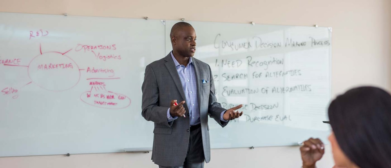 A lecturer speaks to a class while standing in front of a whiteboard