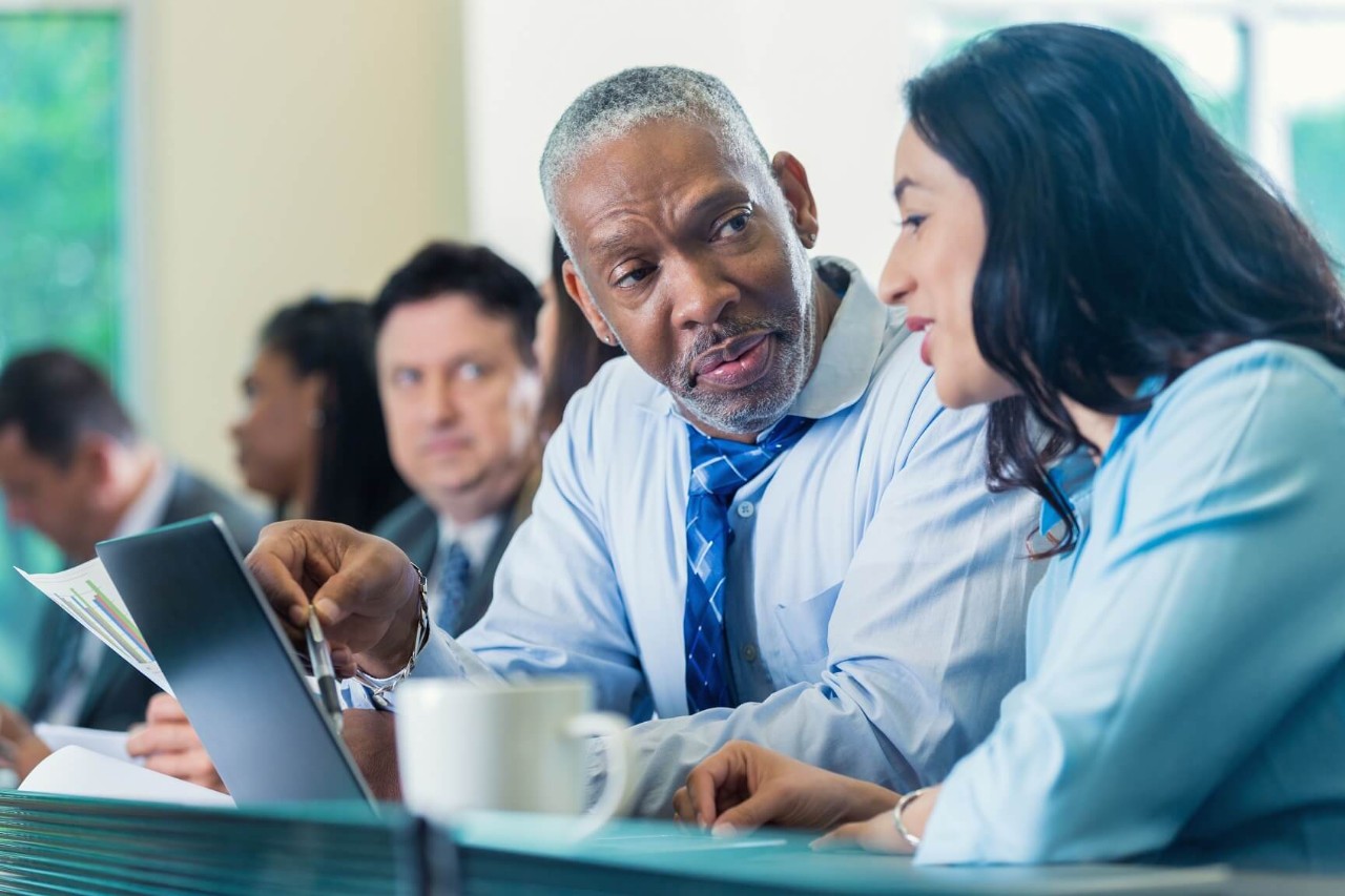 Older male in a group of people sitting at a conference looking at a colleague near him
