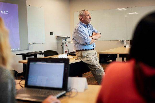A professor leans on a desk during a discussion with students