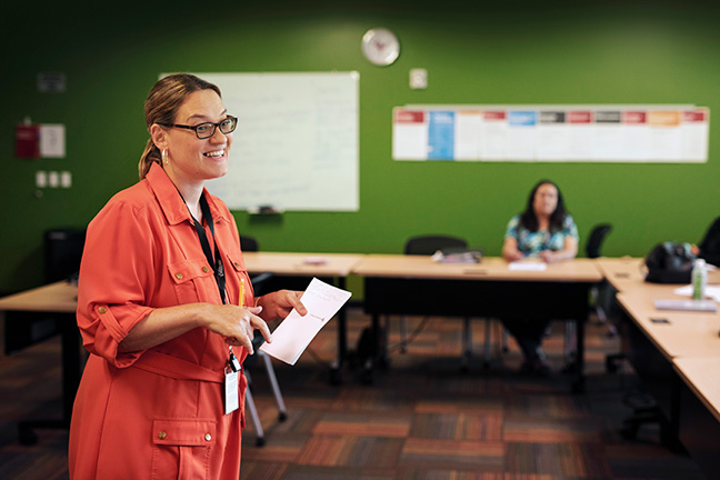 A teacher stands in the middle of a class, smiling and speaking to her students