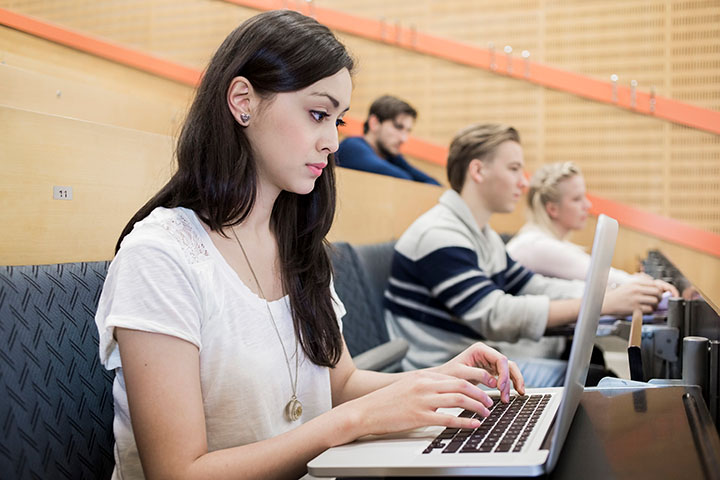 College-aged students sit in a lecture-style hall, listening and taking notes on laptops and tablets